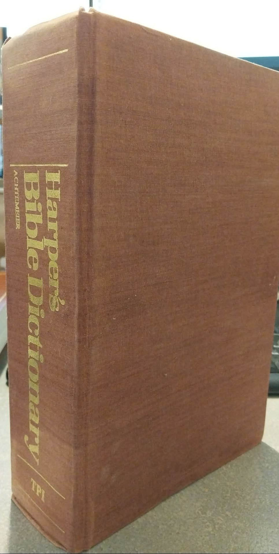 A vintage hardcover dictionary, specifically Harper's Bible Dictionary, Volume 57, shown standing upright on a desk. The book has a textured reddish-brown cloth cover with gold lettering on the spine.