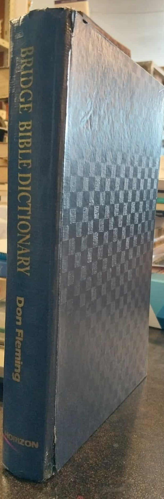 Blue book titled 'Bridge Bible Dictionary' on a cluttered desk with shelves in the background