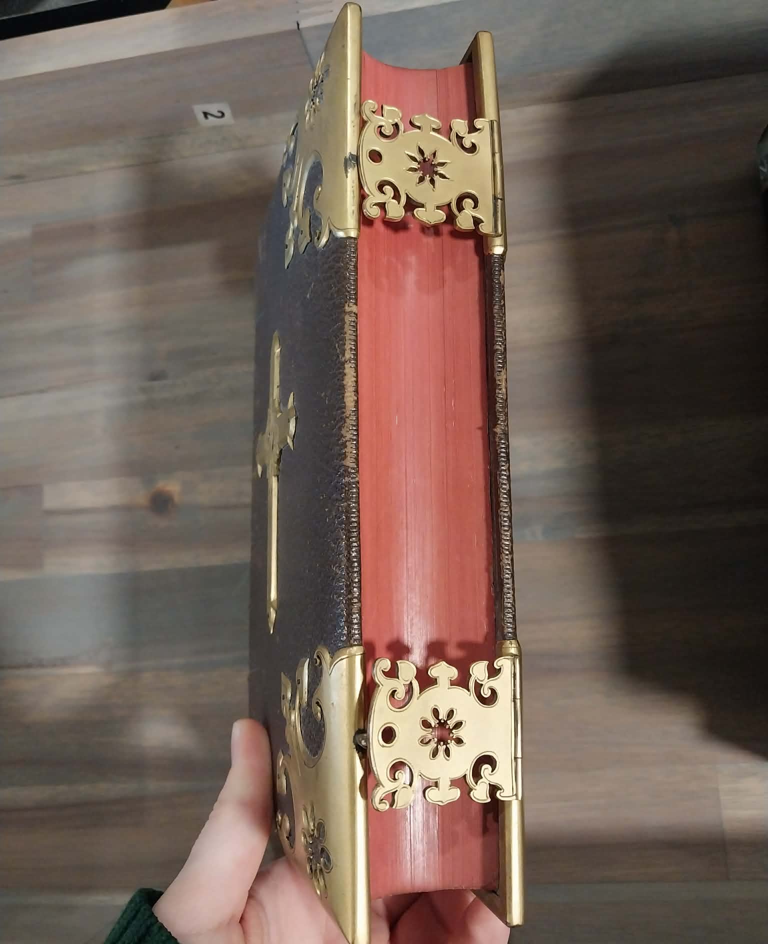 A hand holds a decorative antique-style book with a dark brown textured cover, a pinkish-red spine, and ornate gold metal corner protectors and a cross on the spine, set against a wooden table background.