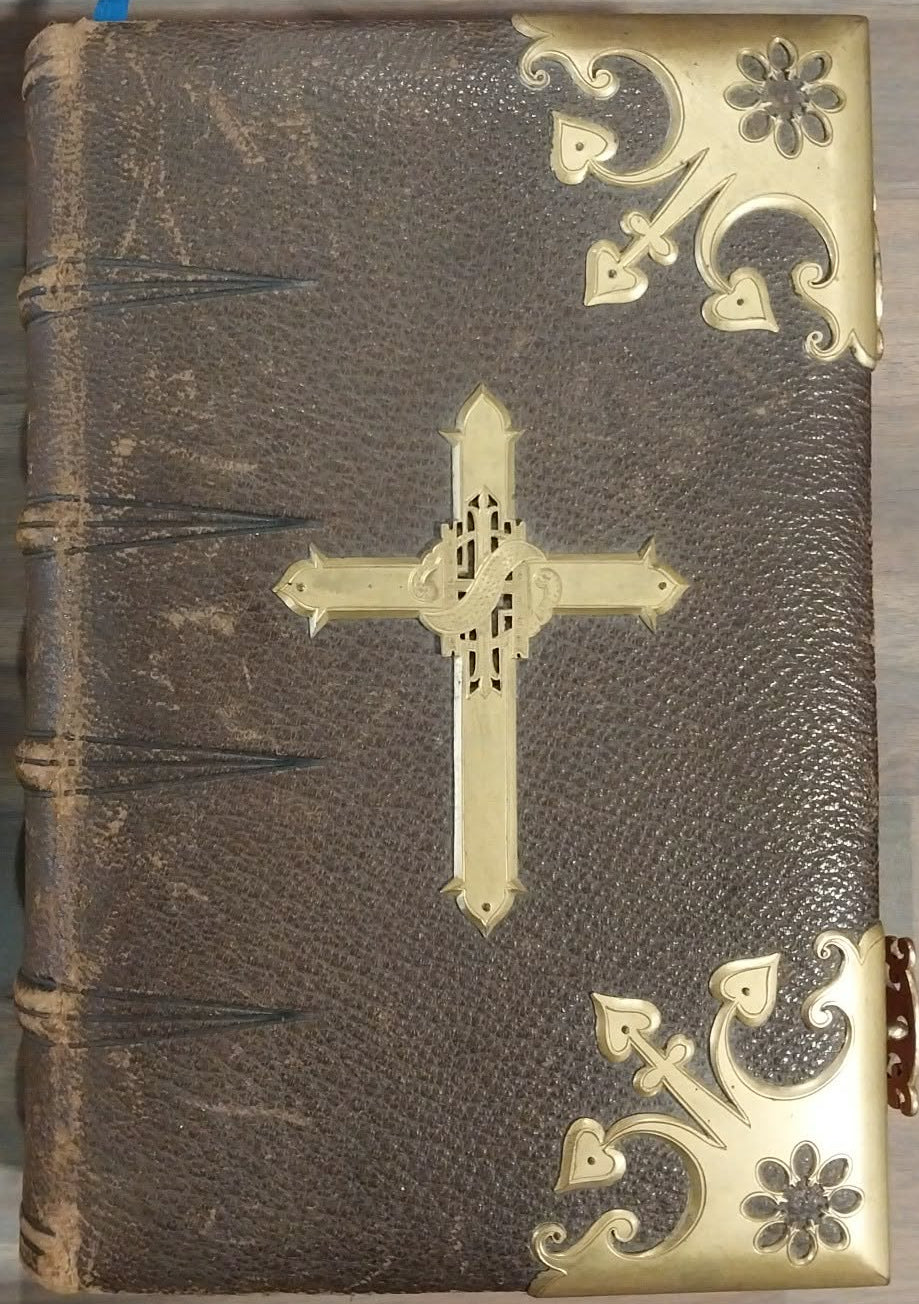 A brown leather-bound book with ornate gold metal corner guards and a central gold cross on the cover, photographed from above on a wood floor.