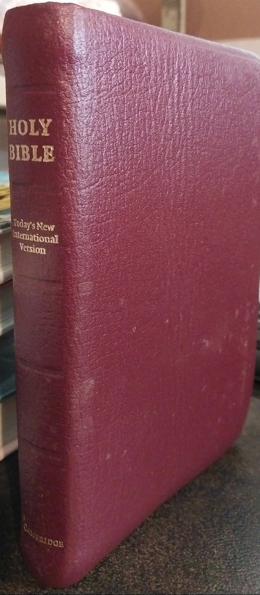 A maroon leather-bound Holy Bible standing upright on a desk, with gold lettering on the spine, photographed in a real indoor setting with a keyboard and shelves in the background.