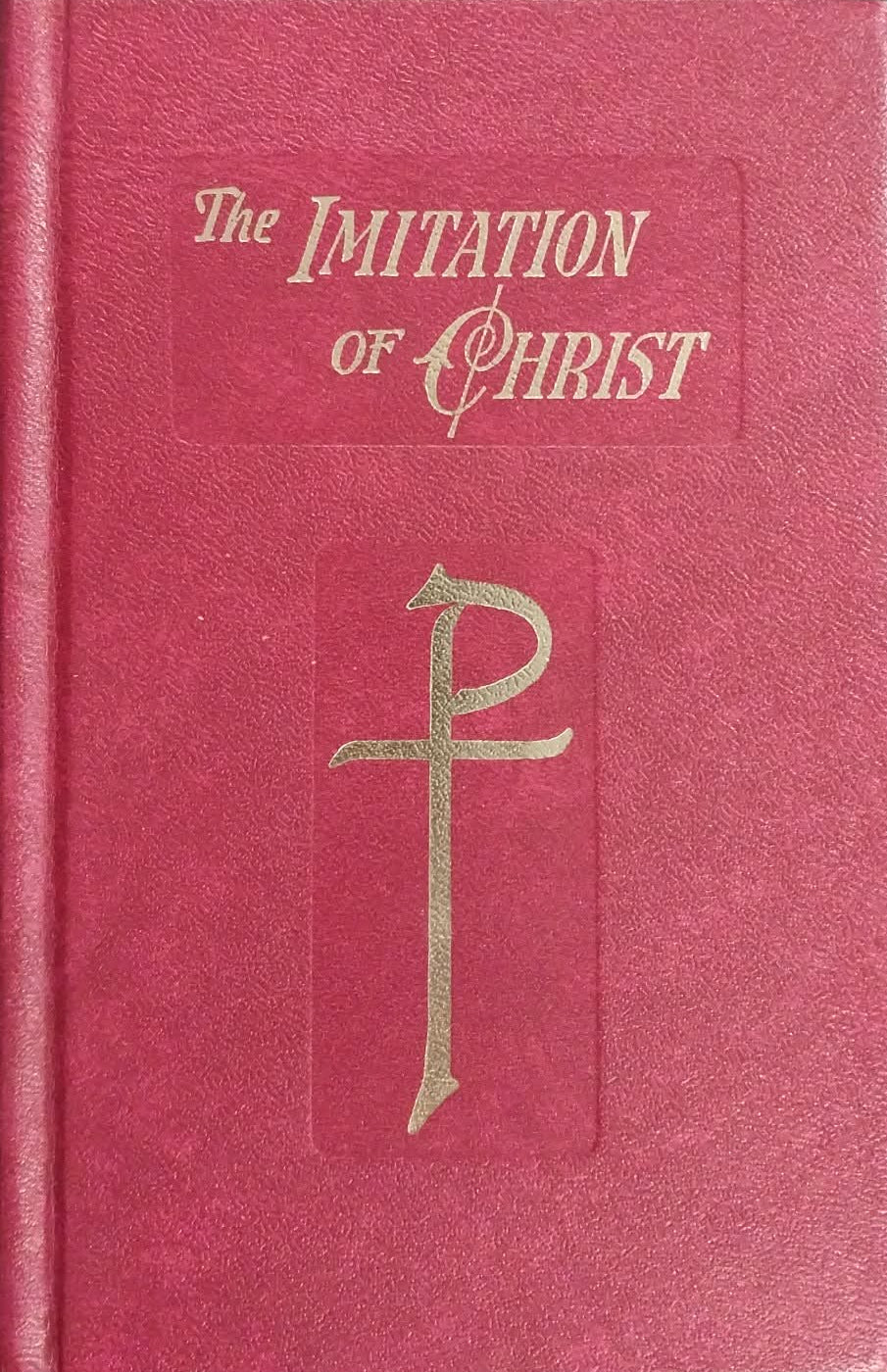 A red hardcover book titled 'The Imitation of Christ' with gold lettering and a gold Chi-Rho symbol on the front cover, photographed on a dark surface.