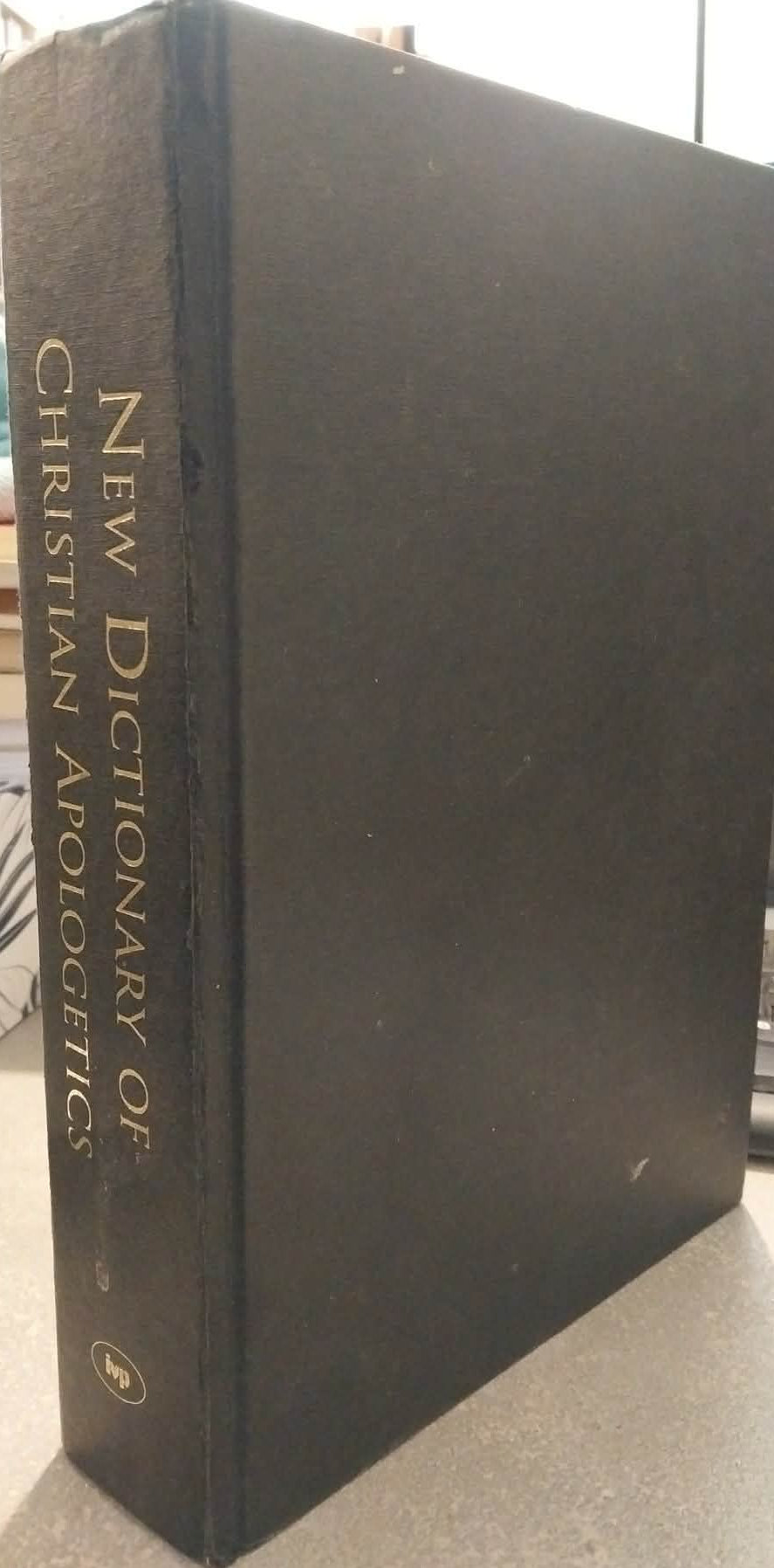 A hardcover book titled 'New Dictionary of Christian Apologetic' standing upright on a desk, with gold lettering on the spine and front cover, photographed in an indoor setting with a blurred keyboard and shelves in the background.