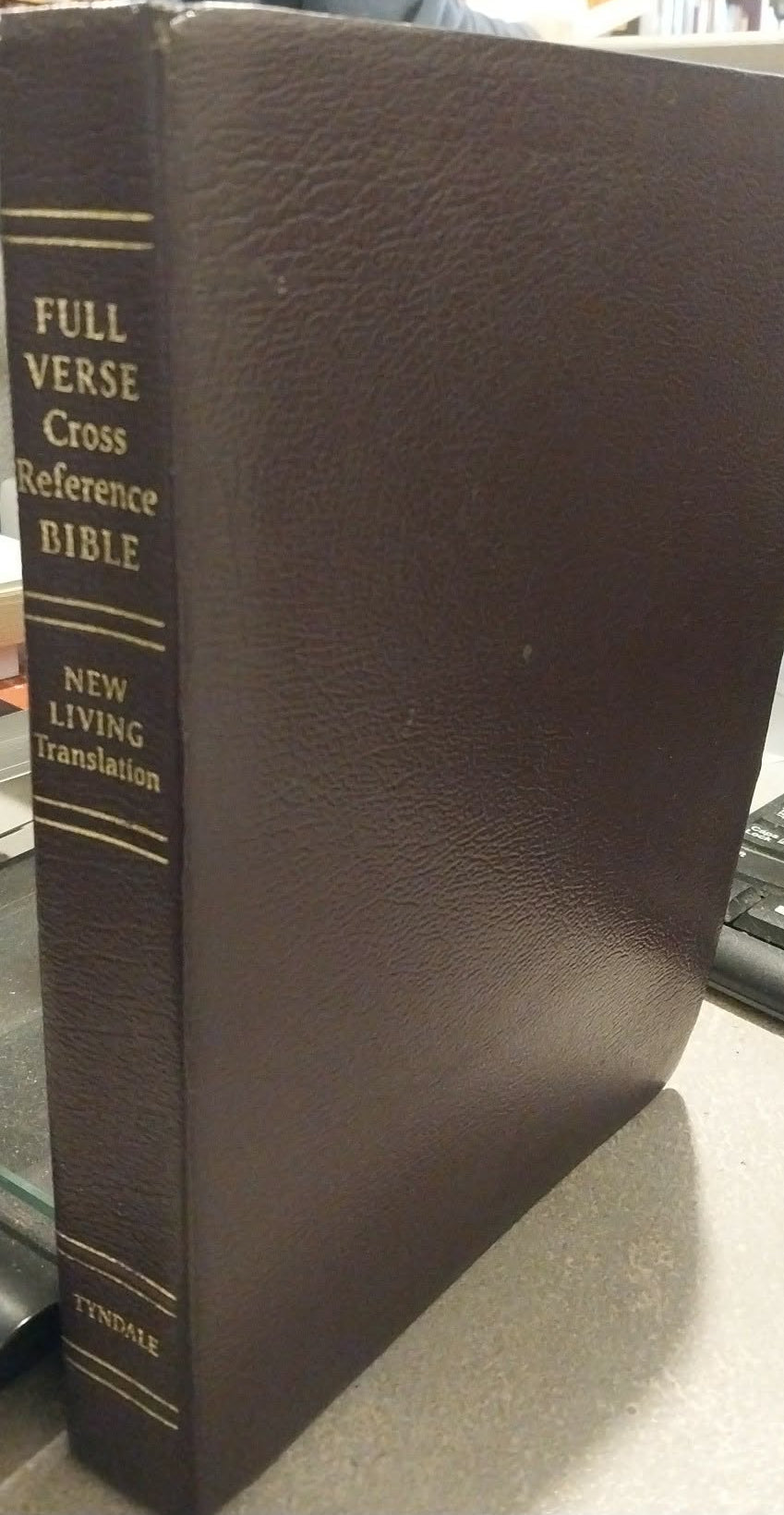 A dark brown leather-bound Bible standing upright on a desk in a library or bookstore, with gold lettering on the spine reading 'FULL VERSE CROSS REFERENCE BIBLE' and 'NEW LIVING Translation.'.