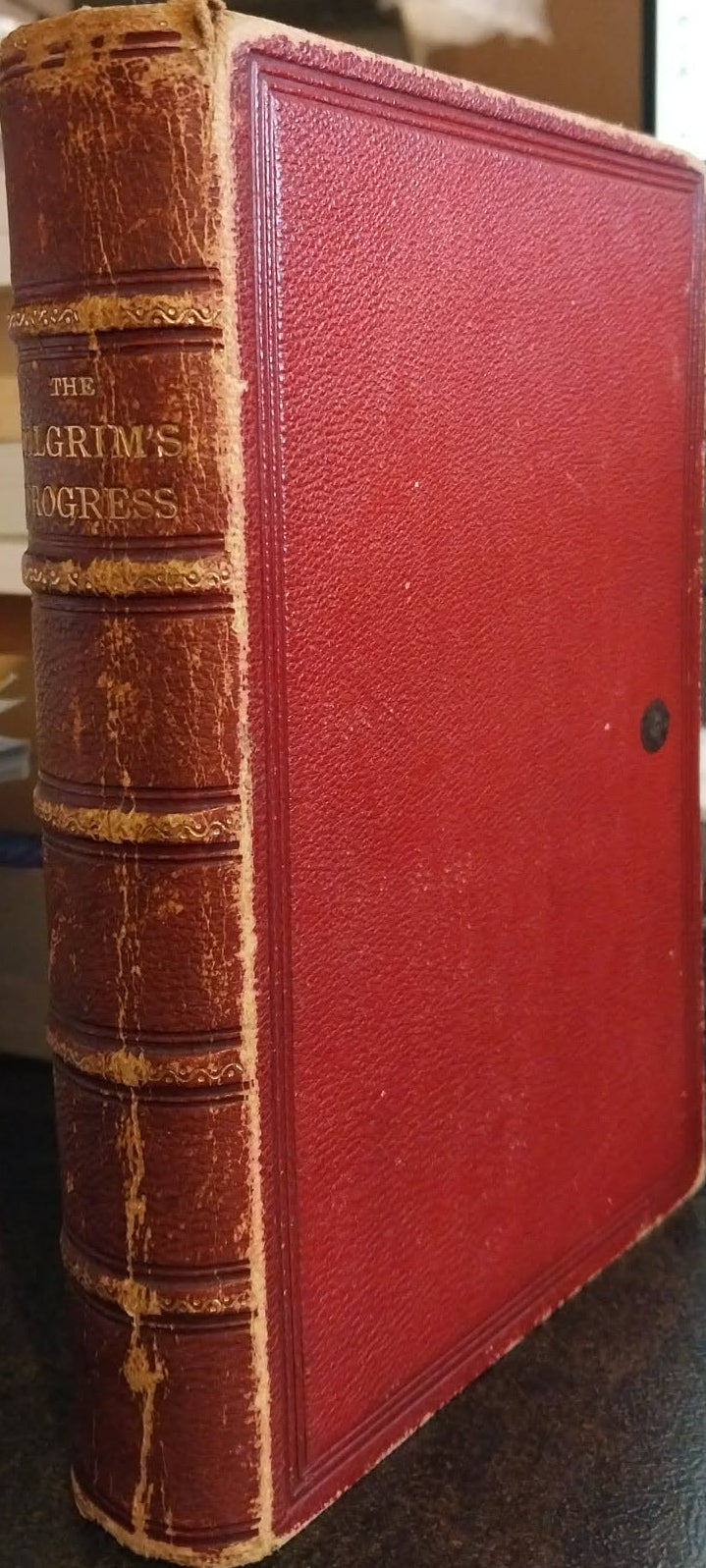 A vintage hardcover book titled 'The Grimness of the Day' with a textured red leather cover and gold-embossed spine detailing, standing on a desk in a cluttered room.