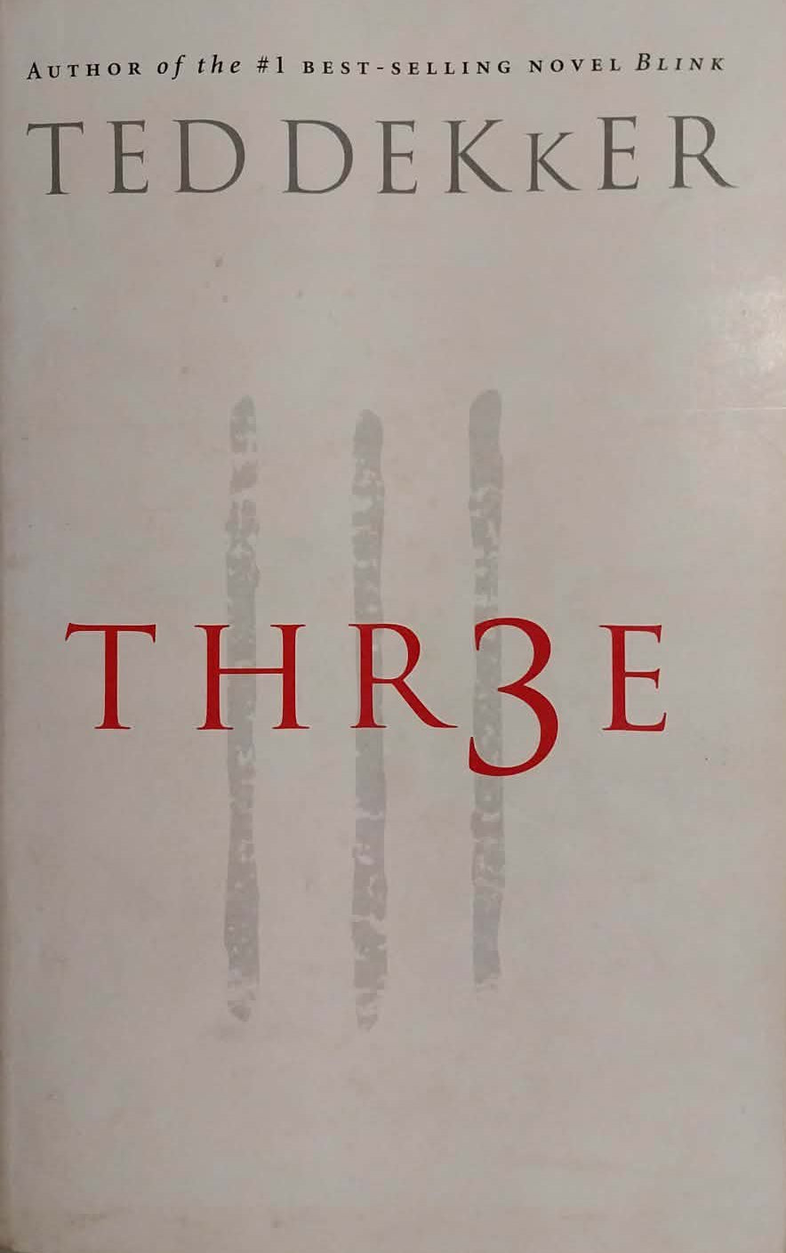 The book cover of 'Thr3e' by Ted Dekker, featuring a white background with red title lettering and gray vertical handprints, photographed on a gray textured surface.
