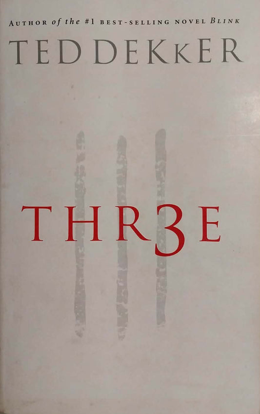 The book cover of 'Thr3e' by Ted Dekker, featuring a white background with red title lettering and gray vertical handprints, photographed on a gray textured surface.